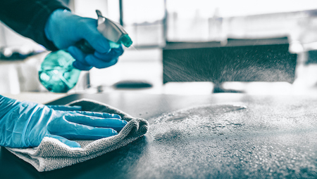 A person wearing gloves sprays a surface with disinfectant and wipes it down with a cloth